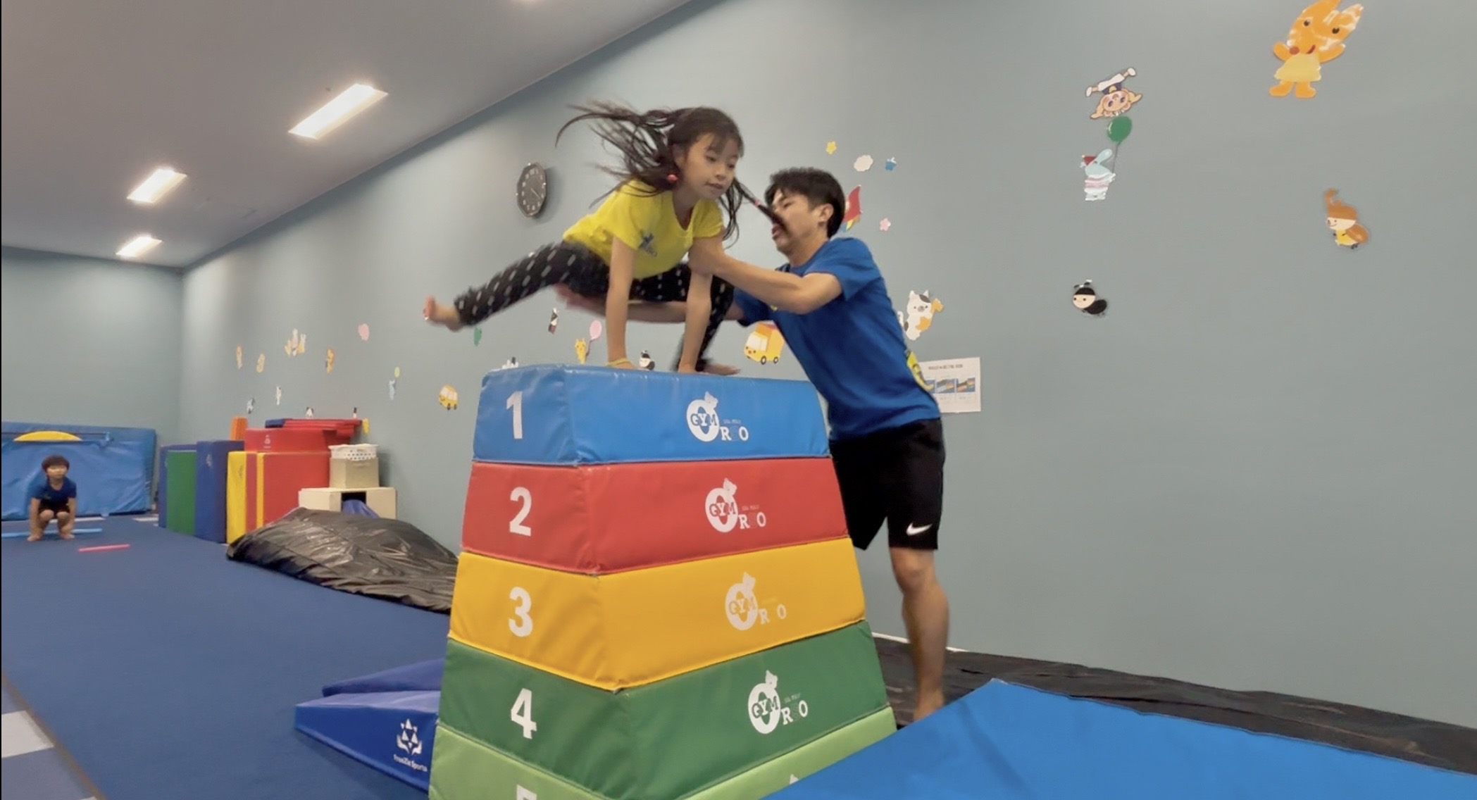 横向き5段の跳び箱を開脚跳びで跳ぶ女の子と補助をする体操教室の先生の様子