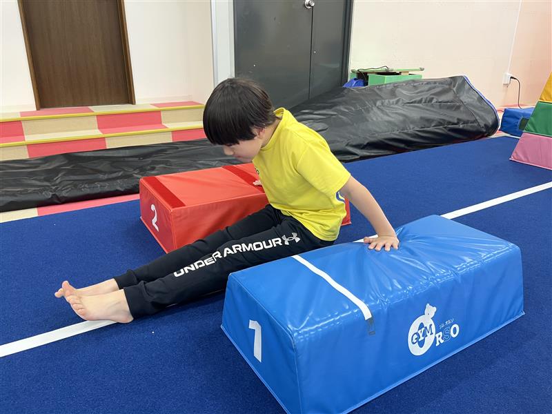 伸膝の状態で補助ブロックを挟み、ブロックを押すことで手で地面を押す感覚を練習する男の子の様子