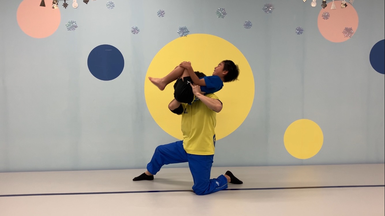 トランポリンで宙返りをする感覚をつかむために、体育座りの体勢で先生に持ち上げられている男の子の様子