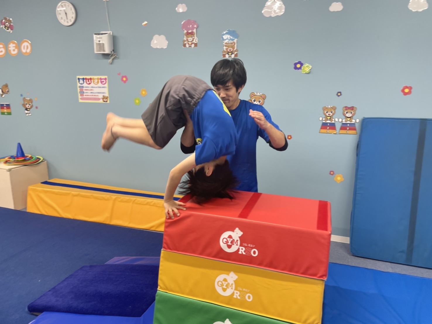 ロイター版で踏切り、頭を跳び箱の上につけて台上前転を行う生徒とサポートする先生の様子