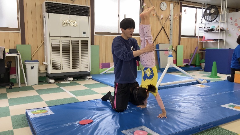 マットの上で倒立をする女の子と支える体操教室の先生の様子