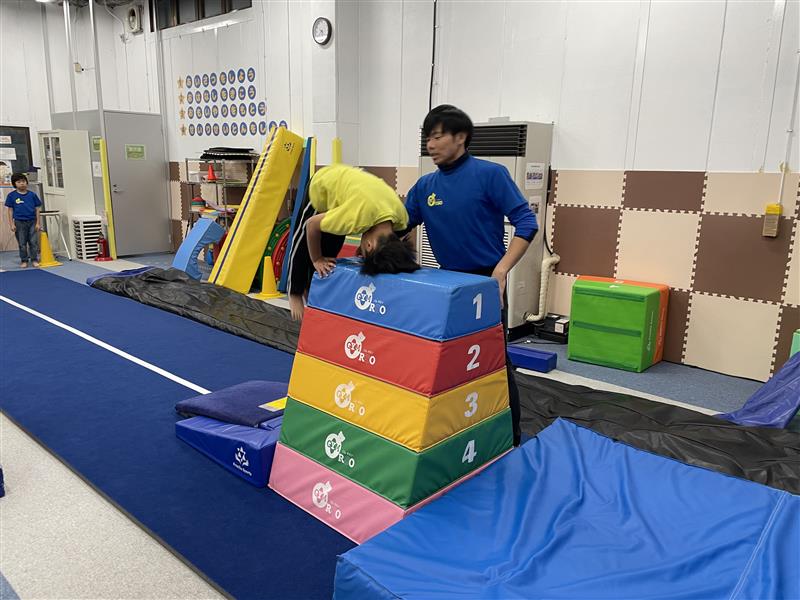 ロイター版で踏切り、膝を伸ばしたまま頭を跳び箱の上につけて頭はね跳びの体勢に入っている体操教室の生徒の様子