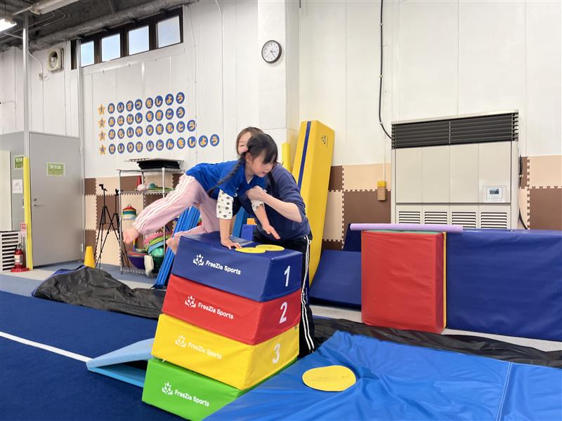 跳び箱の上に乗り、バランスを取りながら四つん這いをしている女の子とそれをサポートしている体操教室の先生の様子