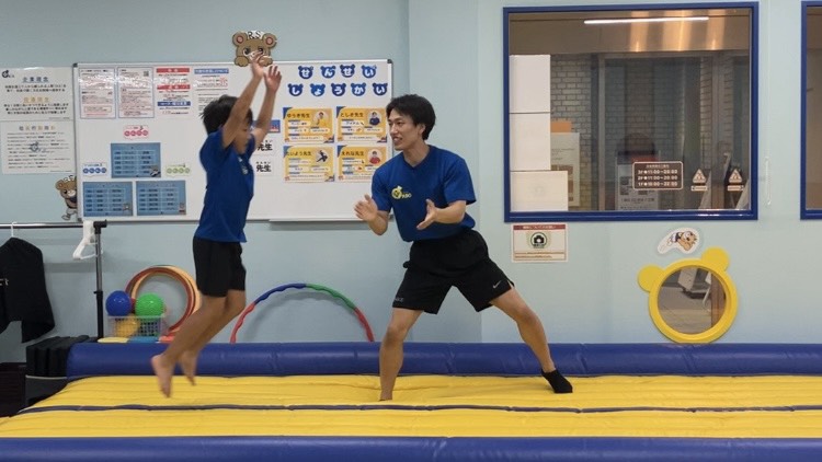 エアータンブリングマットで立ち幅跳びをする生徒と指導する体操教室の先生の様子